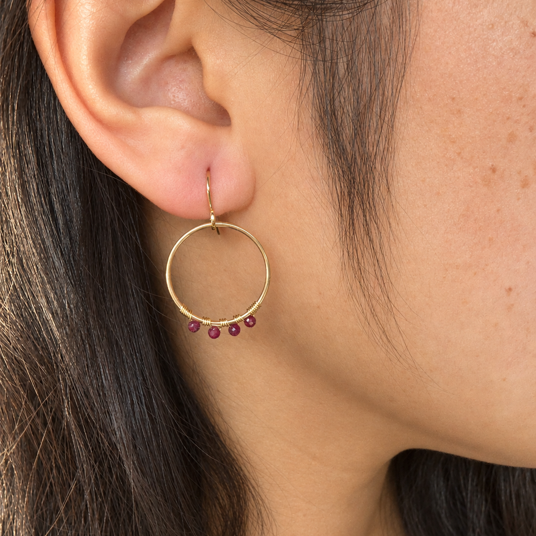 A close-up of a person's ear wearing a delicate gold hoop earring with four small dark red gemstones hanging from the bottom of the hoop.