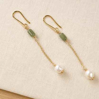A pair of gold dangle earrings with rectangular green stone beads, chain links, and white pearl drops, displayed on a textured beige fabric surface.