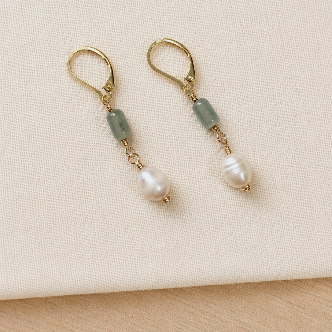 A pair of gold leverback drop earrings featuring a light green stone and a white baroque pearl, displayed on a textured cream fabric background.