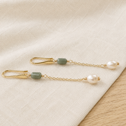 A pair of gold-tone dangle earrings with smooth olive green beads and white pearl drops, displayed on a textured cream linen fabric.