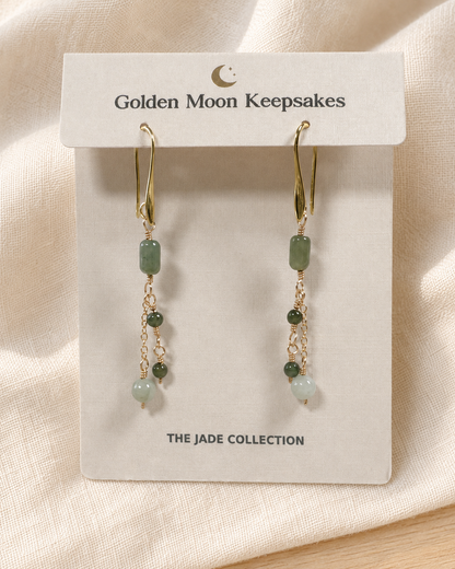 A pair of gold-tone dangle earrings with green jade-like beads, displayed on a white card labeled 'Golden Moon Keepsakes' against a soft beige fabric background.