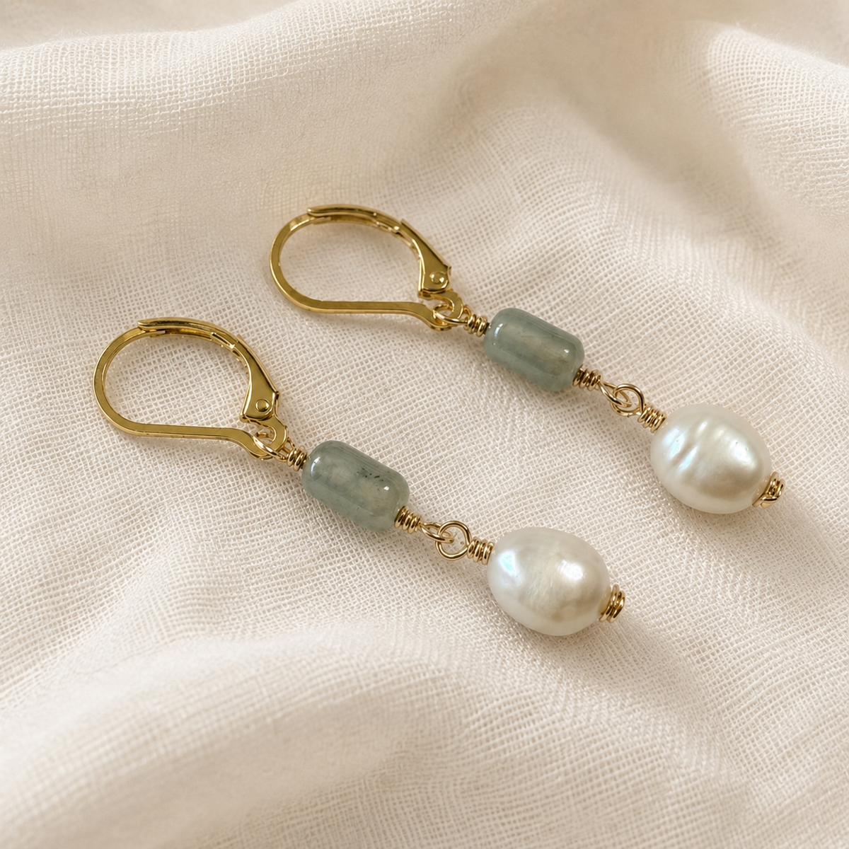 A pair of gold leverback dangle earrings featuring light green translucent beads and white baroque pearls, displayed on a textured cream linen fabric.