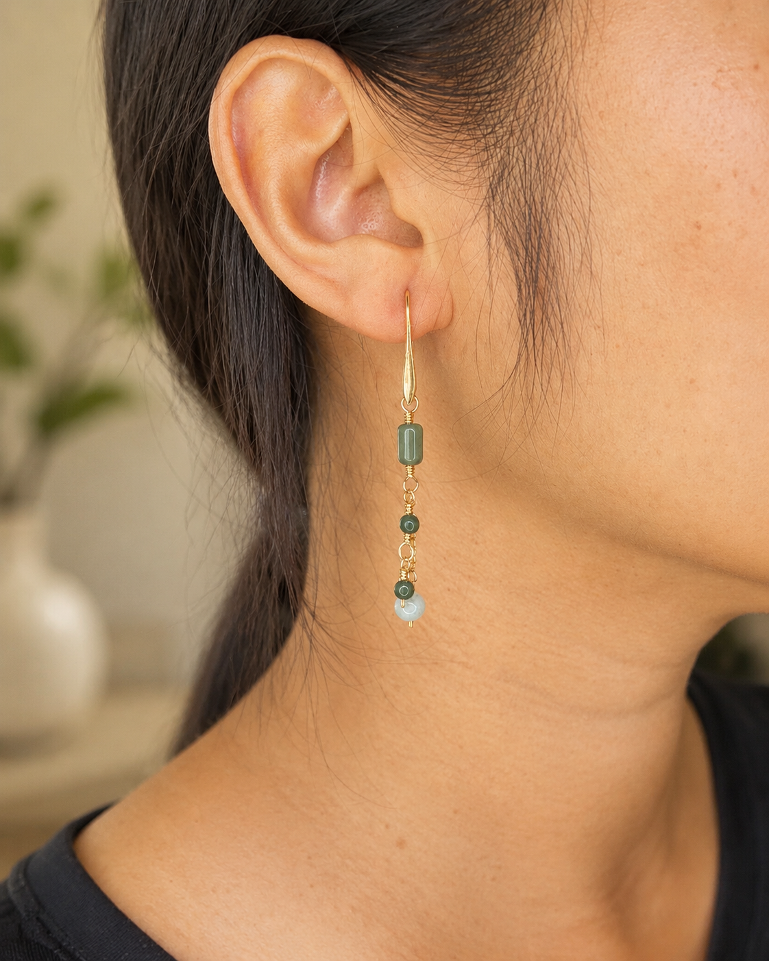 A close-up of a person's ear and neck wearing a long dangle earring. The earring has a gold-toned metal hook, a dark green cylindrical bead, two small dark green beads, and a light blue faceted bead at the bottom.