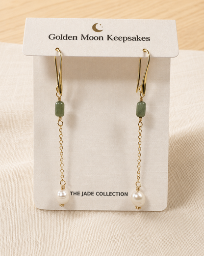 A pair of gold dangle earrings with green jade-like beads and white pearl drops, displayed on a white card labeled 'Golden Moon Keepsakes' and 'THE JADE COLLECTION'.