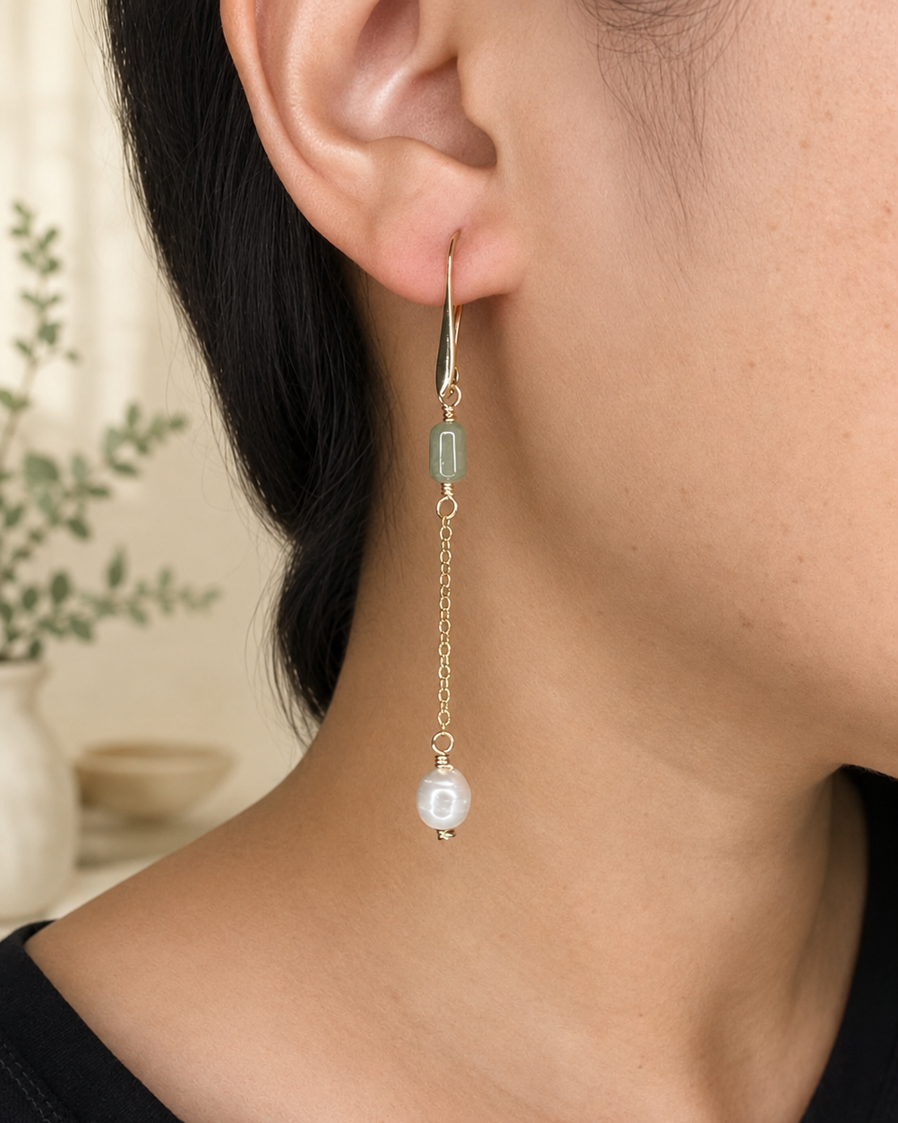 A close-up of a person's ear wearing a delicate gold dangle earring with a light green stone, a small gold chain, and a white pearl bead.