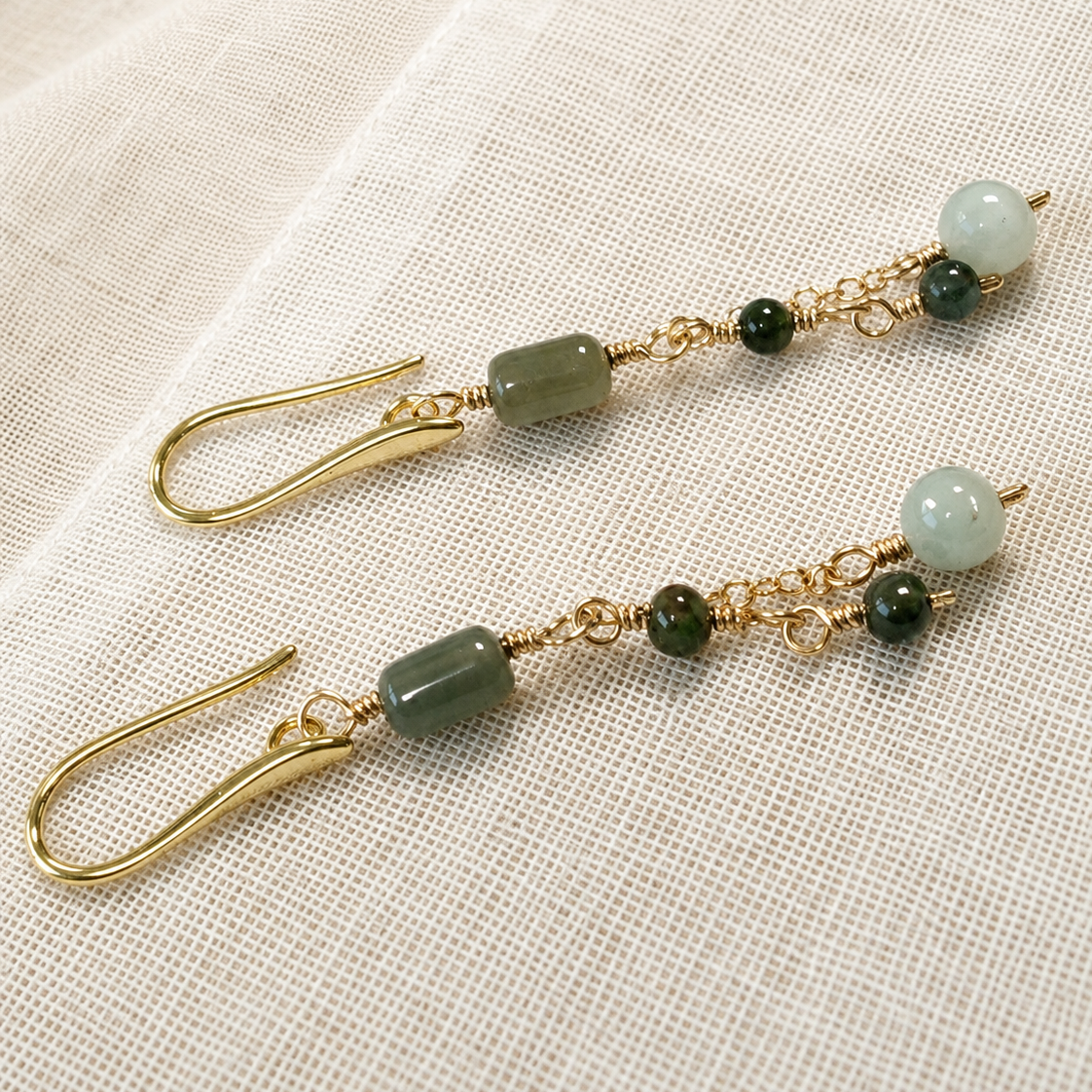 A pair of gold-tone dangle earrings with green stone beads and small dark green accent beads, displayed on a textured cream linen fabric background.