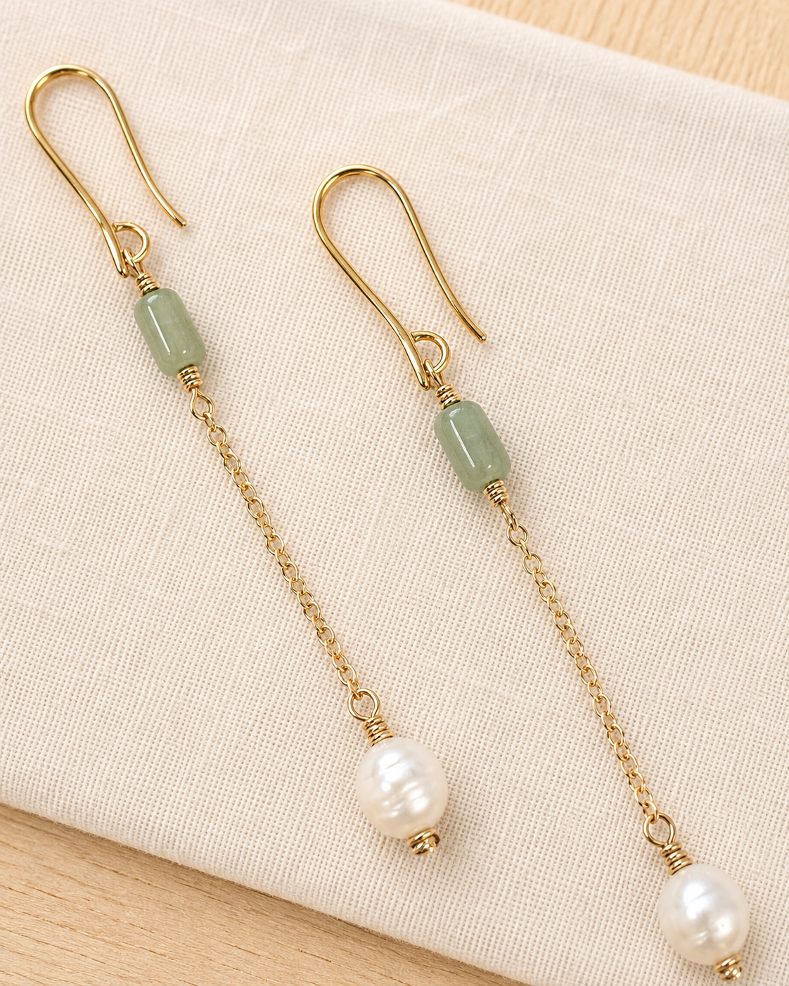 A pair of gold-tone dangle earrings with light green stone beads and white pearl drops, displayed on a textured cream fabric background.