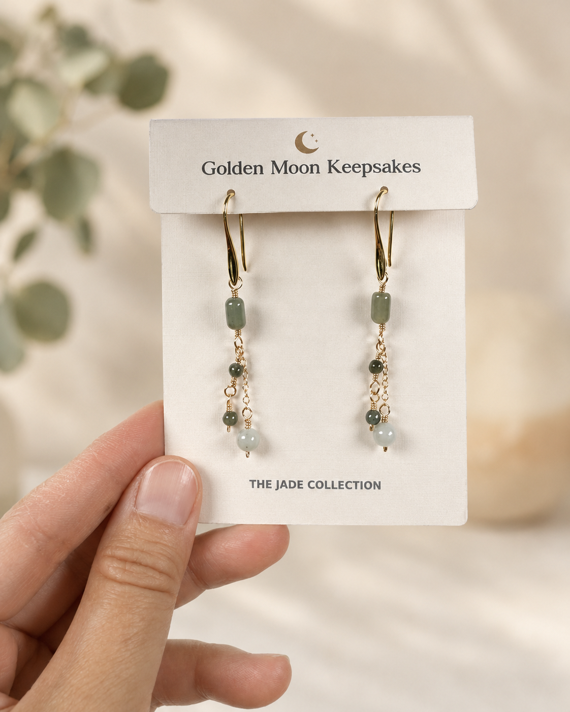 A hand holds a white display card featuring a pair of dangling earrings from Golden Moon Keepsakes. The earrings have gold-tone hooks and chains with small green and pale blue beads.