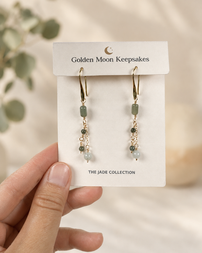 A hand holds a white display card featuring a pair of dangling earrings from Golden Moon Keepsakes. The earrings have gold-tone hooks and chains with small green and pale blue beads.