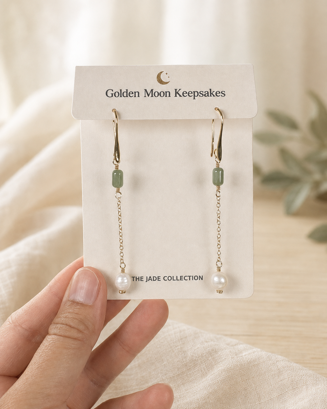 A pair of gold drop earrings with light green rectangular beads and white pearl-like beads, displayed on a white card labeled 'Golden Moon Keepsakes' and held by a hand against a soft neutral background.