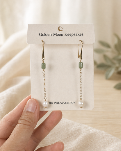 A pair of gold drop earrings with light green rectangular beads and white pearl-like beads, displayed on a white card labeled 'Golden Moon Keepsakes' and held by a hand against a soft neutral background.