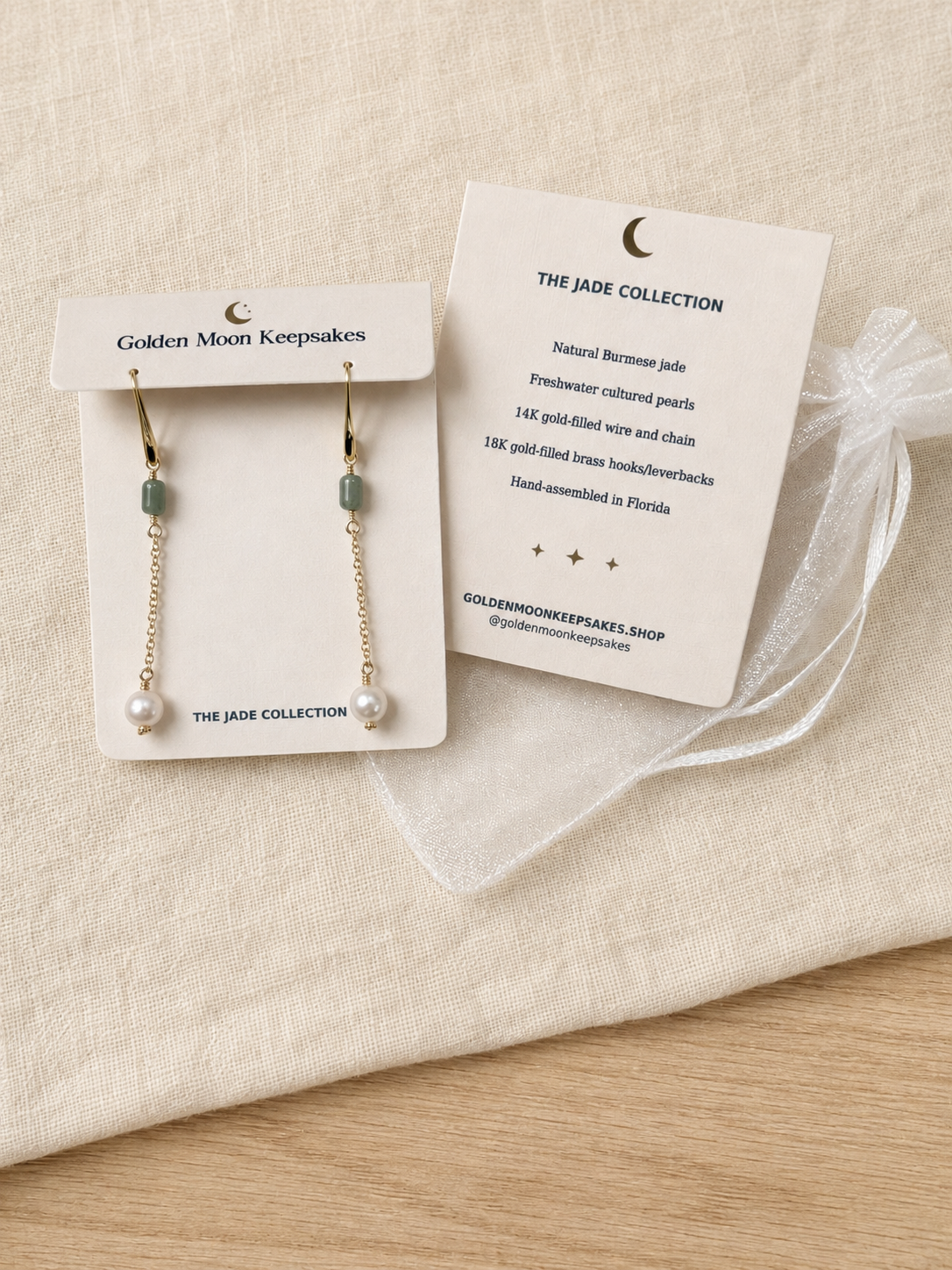 A pair of gold dangle earrings with green jade-like beads and white pearl drops, displayed on two white cards from The Jade Collection on a beige fabric surface.