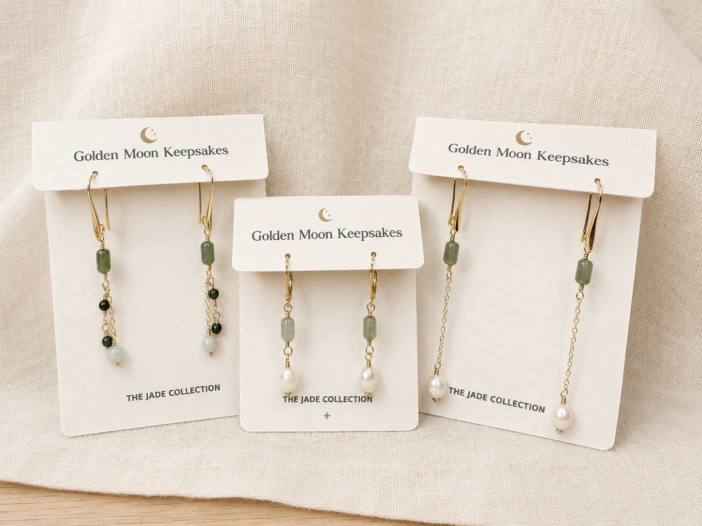 Three pairs of dangle earrings displayed on white card holders labeled Golden Moon Keepsakes, arranged on a light beige fabric background. The earrings feature gold-tone hooks and chains with pale green rectangular beads, small dark beads, and white pearl-like drops.