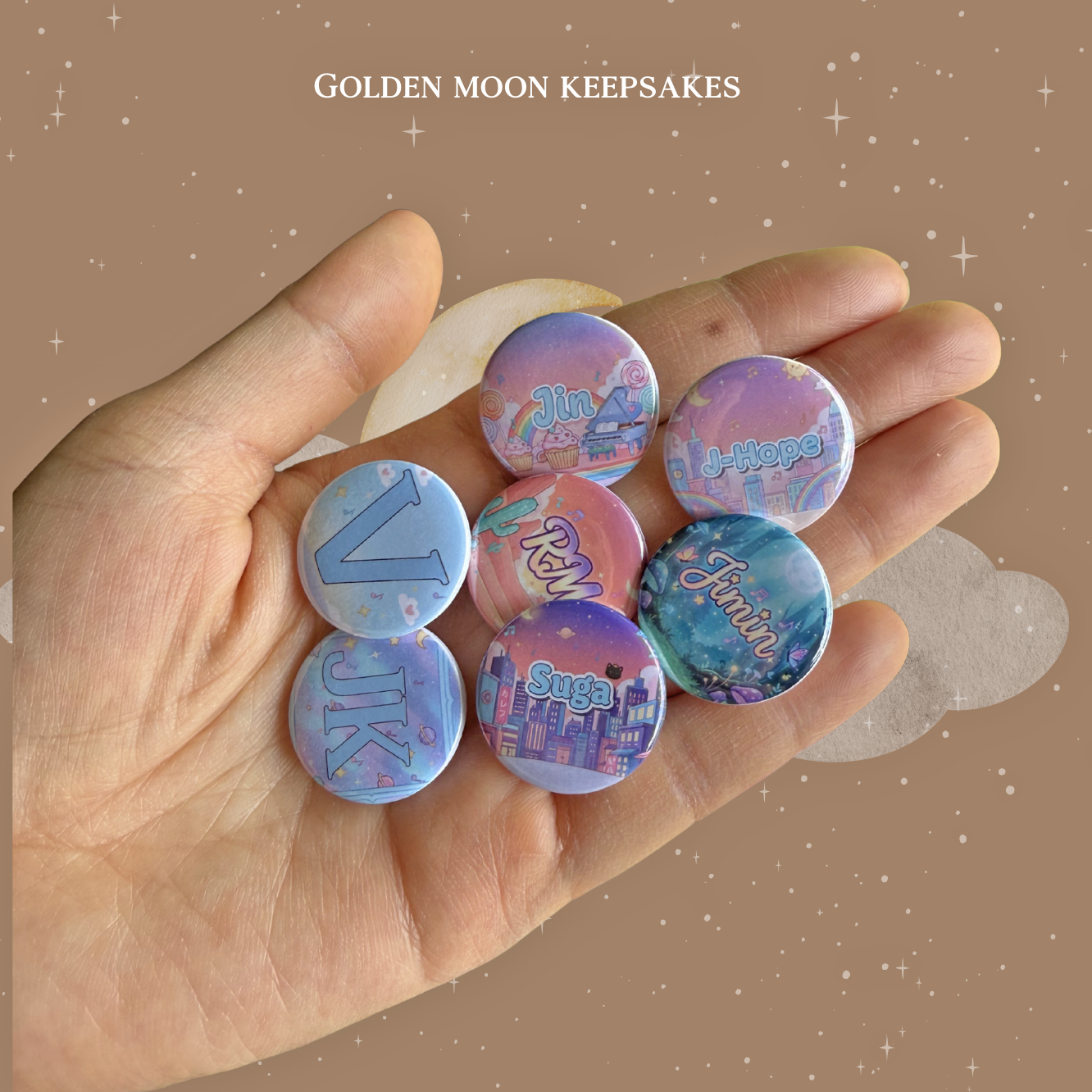 A hand holding seven 1-inch round button pins, each featuring a different name or set of initials: Jin, J-Hope, V, RM, Jimin, JK, and Suga. The names are written in stylized, colorful fonts over various themed backgrounds like cityscapes, rainbows, and galaxy patterns. The image is presented on a tan celestial-themed background with the shop name at the top.