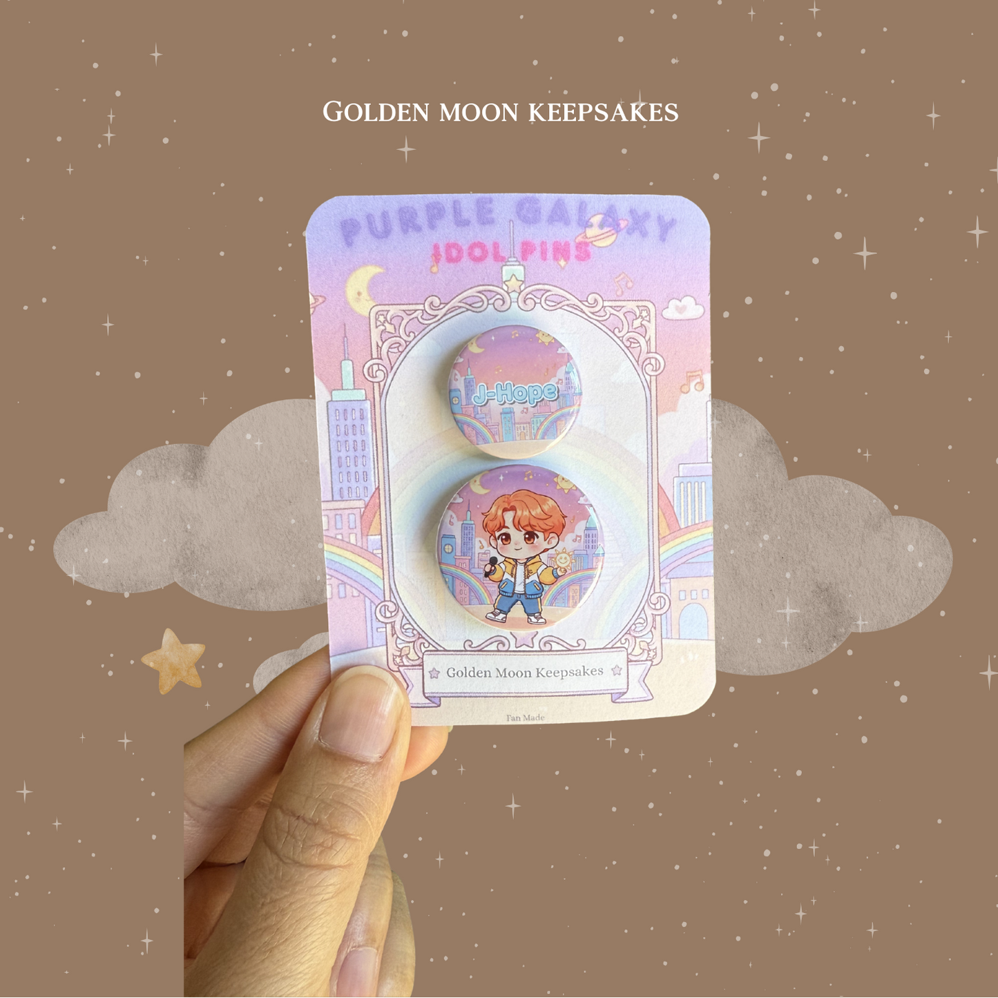 A hand holding a custom 3x4 inch backing card for "Golden Moon Keepsakes" featuring a two-piece pin set. The card displays a 1-inch pin with the name "J-Hope" and a 1.25-inch pin with a matching chibi character. Both pins feature a vibrant cityscape and rainbow background under a purple and pink sky.