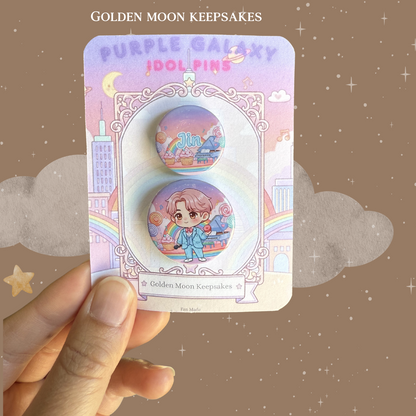 A hand holding a 3x4 inch "Golden Moon Keepsakes" backing card with a two-piece button pin set. The set includes a 1.25-inch pin with a chibi character in a blue suit and a 1-inch pin with the name "Jin". The design features a whimsical "Idol World" background with rainbows, cupcakes, and a piano.
