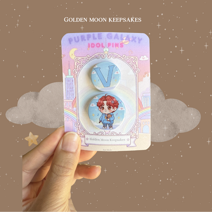 A hand holding a custom "Golden Moon Keepsakes" backing card featuring a two-piece button pin set. The top 1-inch pin displays a large light blue letter "V" against a soft blue sky background with clouds. The bottom 1.25-inch pin features a chibi character with red hair wearing a blue hoodie and holding a small brown bear.