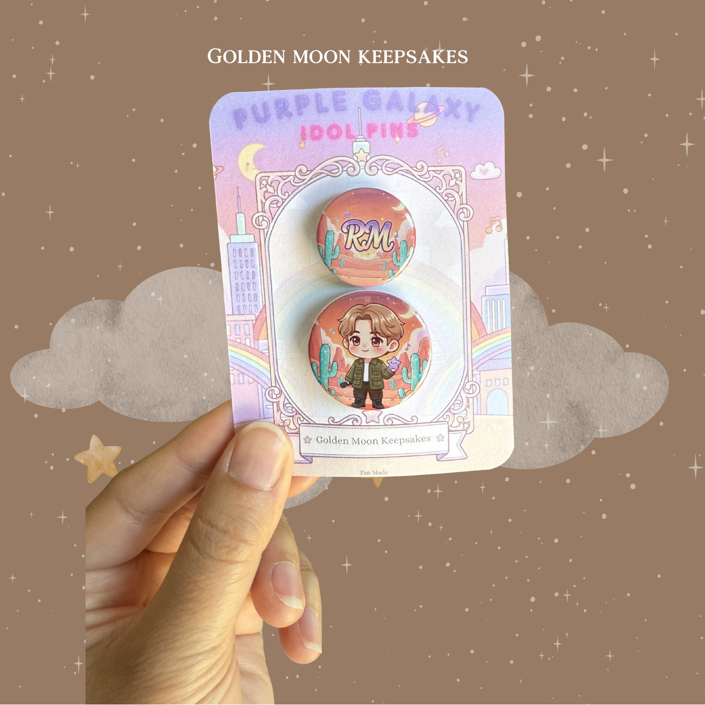 A hand holding a 3x4 inch "Golden Moon Keepsakes" backing card with a two-piece button pin set. The 1.25-inch pin features a chibi character in an olive jacket against an outdoor desert background with cacti. The matching 1-inch pin displays the name "RM" in a stylized font over a similar sunset-toned desert landscape.
