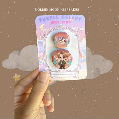 A hand holding a 3x4 inch "Golden Moon Keepsakes" backing card with a two-piece button pin set. The 1.25-inch pin features a chibi character in an olive jacket against an outdoor desert background with cacti. The matching 1-inch pin displays the name "RM" in a stylized font over a similar sunset-toned desert landscape.
