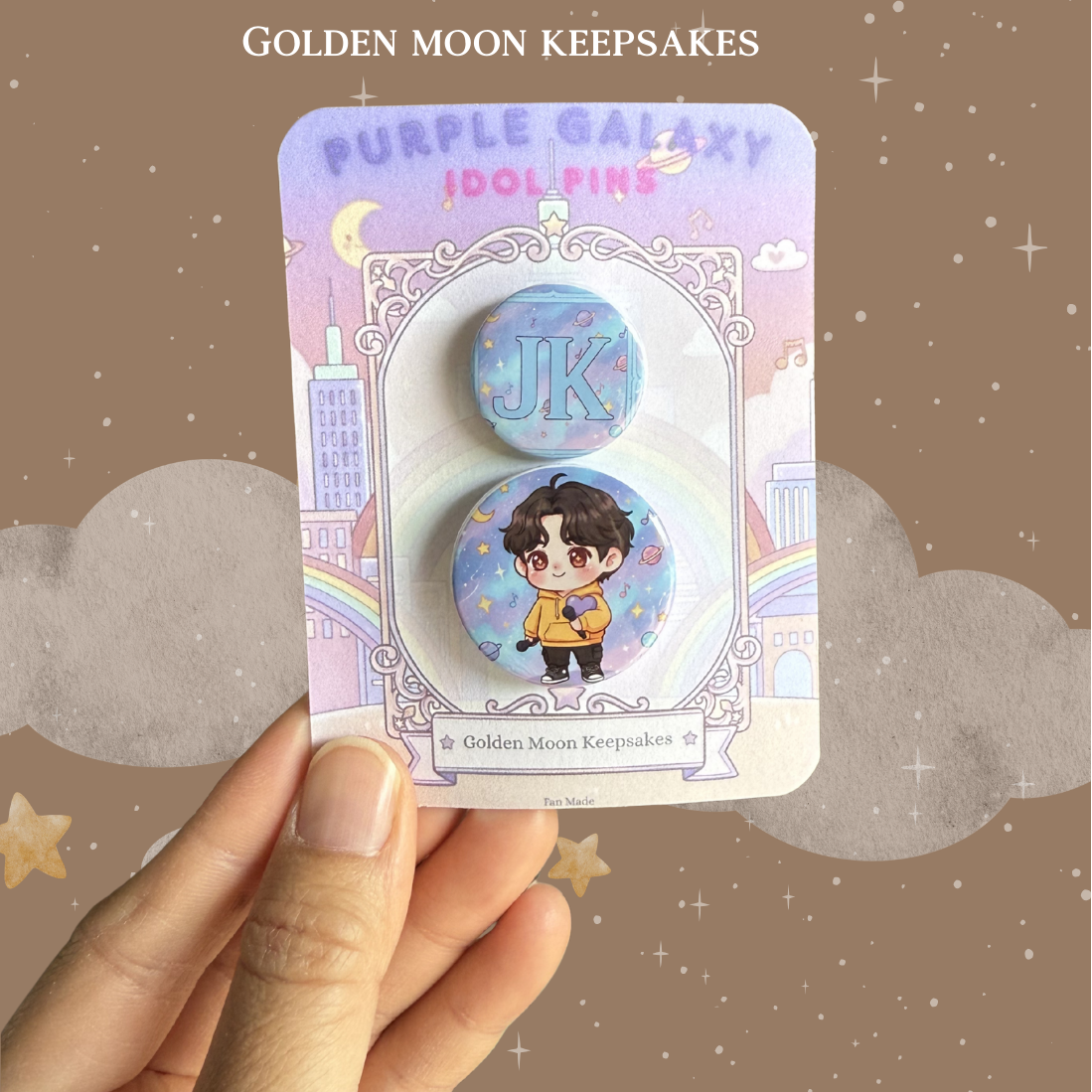 A hand holding a 3x4 inch backing card with a two-piece "Idol World" pin set. The larger pin shows a chibi character in a yellow hoodie holding a purple heart against a starry purple sky. The matching 1-inch pin displays the initials "JK" in a bold blue font over a coordinating celestial background.