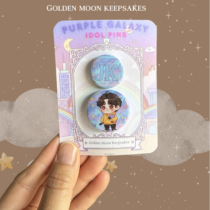 A hand holding a 3x4 inch backing card with a two-piece "Idol World" pin set. The larger pin shows a chibi character in a yellow hoodie holding a purple heart against a starry purple sky. The matching 1-inch pin displays the initials "JK" in a bold blue font over a coordinating celestial background.