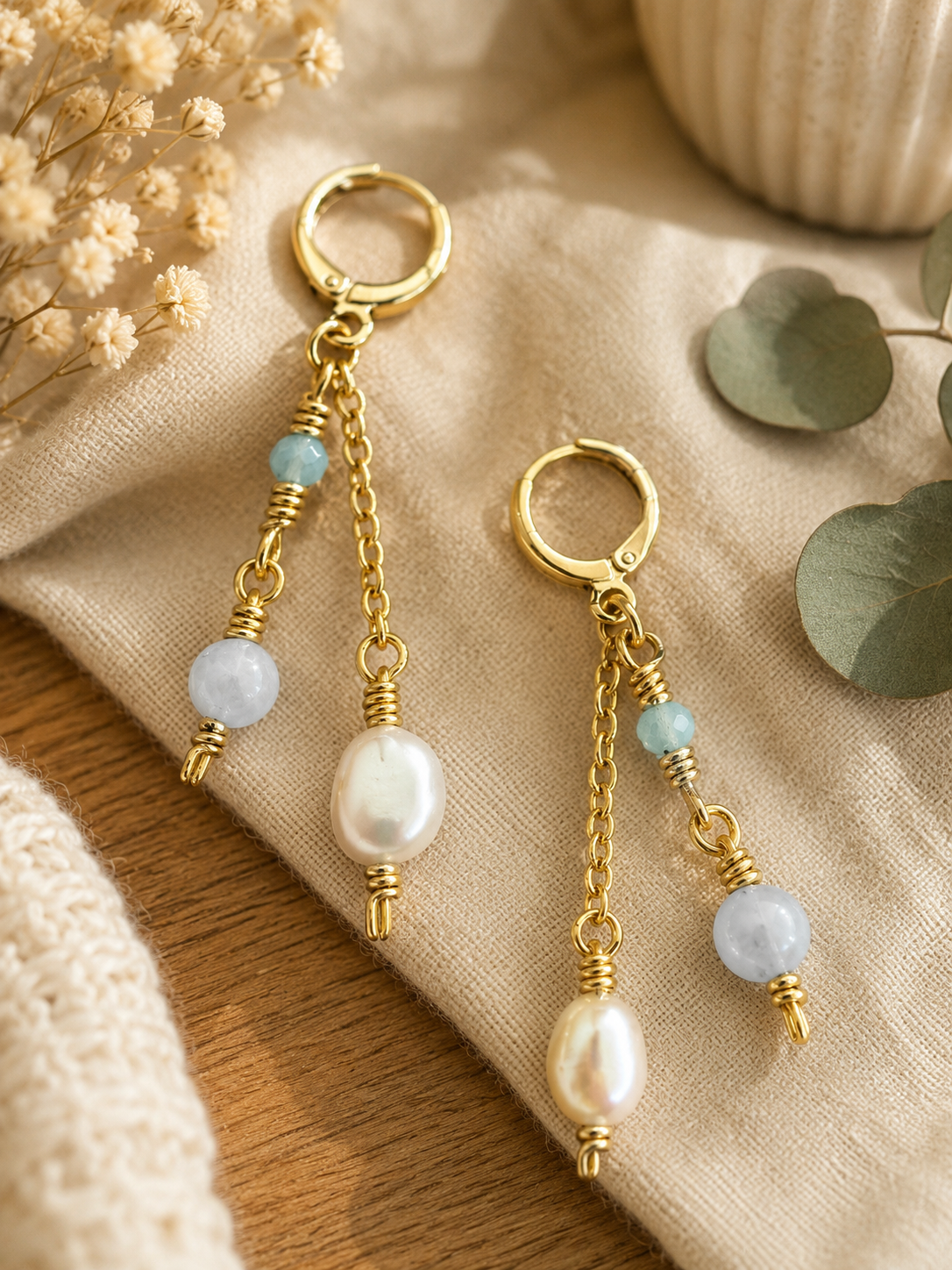 A pair of gold-toned dangle earrings with freshwater pearl drops, light blue beads, and textured wire details, styled on a beige linen cloth with dried flowers and eucalyptus leaves.