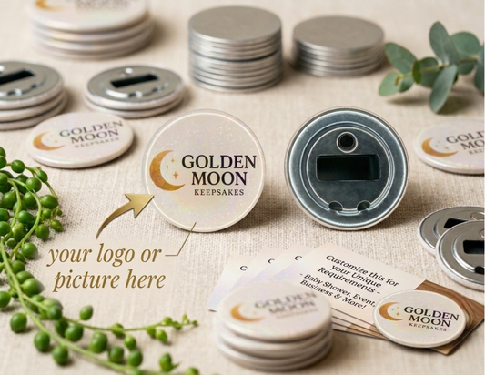 A product mockup showing two circular bottle openers on a light-grey, textured surface: one with a customizable holographic 'Golden Moon Keepsakes' logo and a gold arrow pointing to it, with the text 'your logo or picture here,' and the other showing the metal reverse with the opener and magnet. The background includes stacks of metal blanks, scattered logo openers, and informational business cards, with green plants and natural, soft lighting.