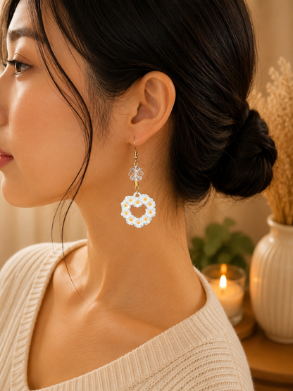 A woman in profile wearing a dangling earring with a white flower ring and a clear faceted bead above it, photographed in a softly lit indoor setting with a candle and dried flowers in the background.