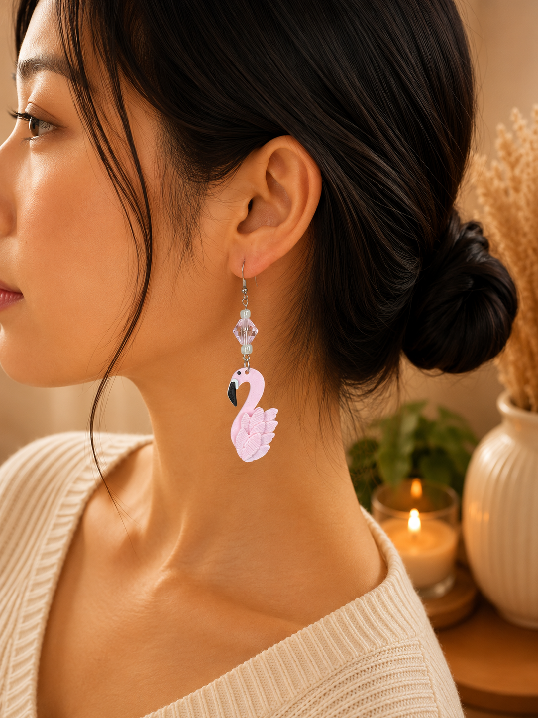 A close-up side profile of a woman wearing a dangling flamingo earring. The earring features a light pink flamingo charm with a black beak, suspended from clear faceted beads.