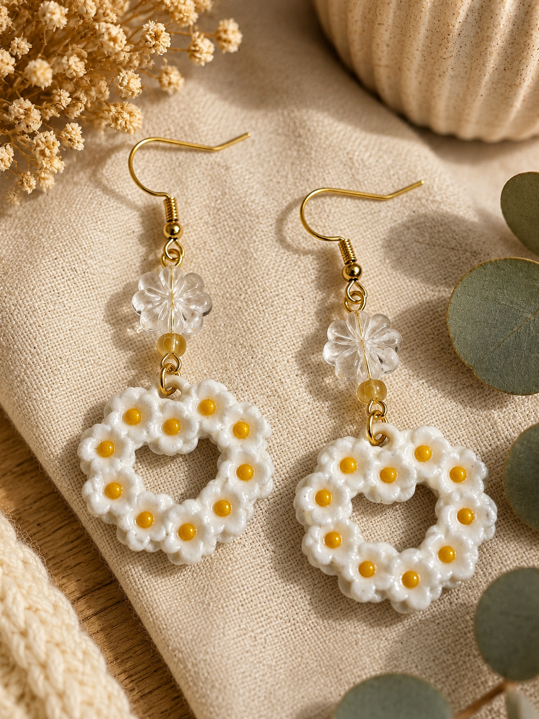 A pair of handmade daisy drop earrings with white textured petals, small yellow bead centers, and gold-tone fishhook wires, displayed on a beige linen fabric with dried flowers and eucalyptus leaves.