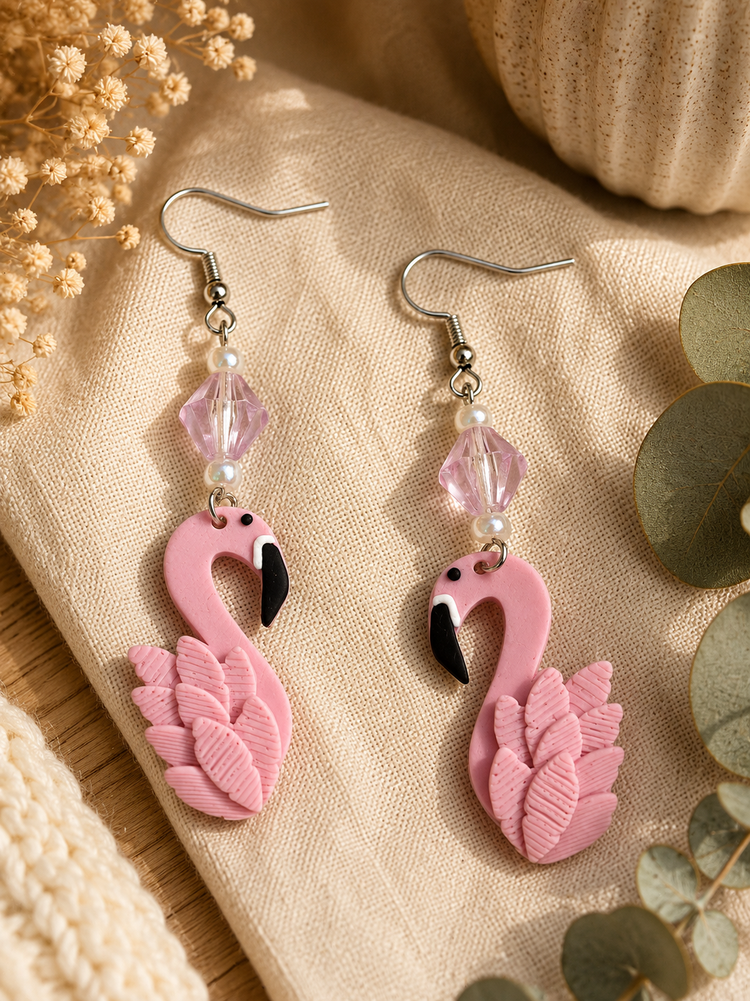 A pair of pink flamingo dangle earrings with silver hooks, clear faceted beads, and small pearl-like accents, displayed on beige fabric with dried flowers and eucalyptus leaves.