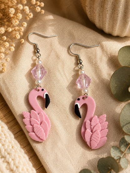 A pair of pink flamingo dangle earrings with silver hooks, clear faceted beads, and small pearl-like accents, displayed on beige fabric with dried flowers and eucalyptus leaves.