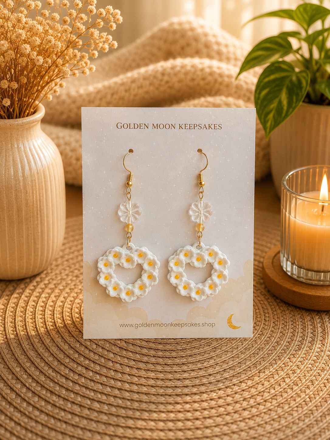A pair of white daisy earrings with yellow centers and gold hooks, displayed on a white card labeled 'GOLDEN MOON KEEPSAKES' on a woven surface with dried flowers, a candle, and a plant in the background.