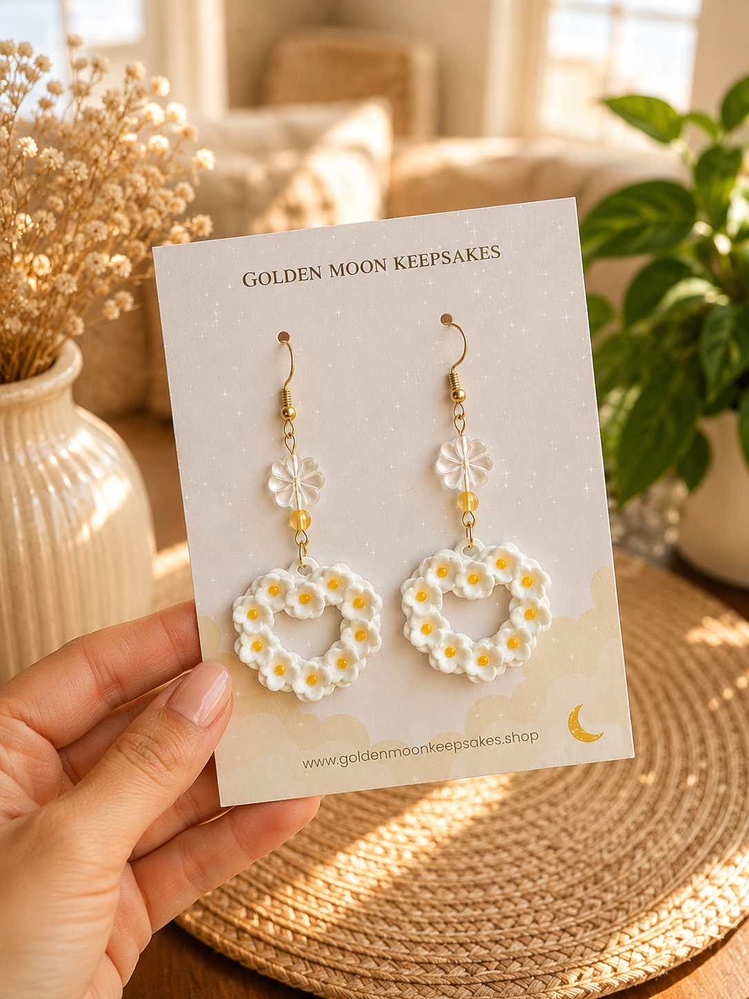 A hand holds a display card featuring a pair of daisy-patterned hoop earrings with gold hooks. The card is labeled 'GOLDEN MOON KEEPSAKES' and is photographed in a warm, sunlit indoor setting with dried flowers and a woven mat in the background.