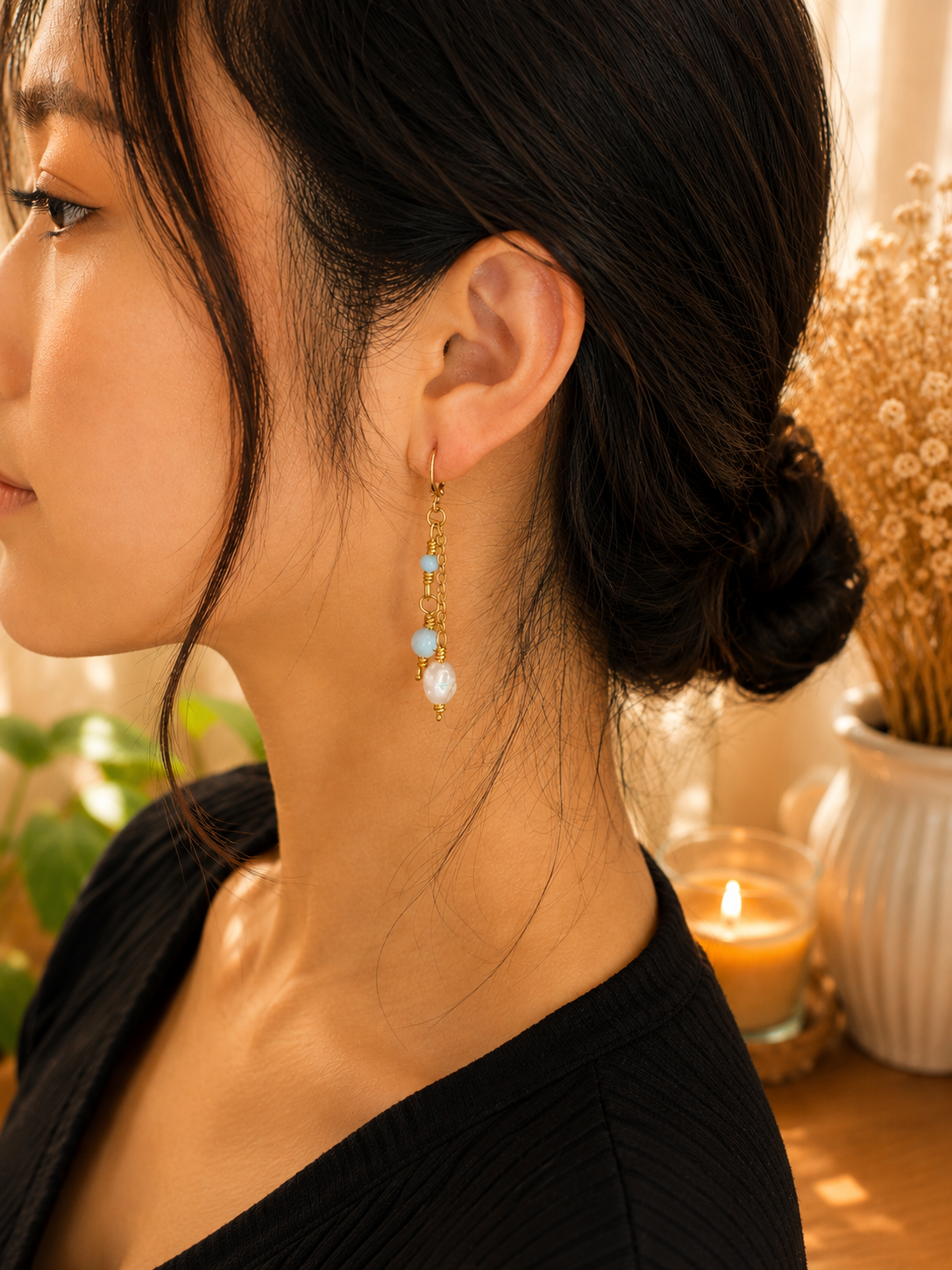 A close-up side profile of a woman wearing a delicate gold dangle earring with light blue beads and a white pearl, set against a warm, softly lit background with a lit candle.