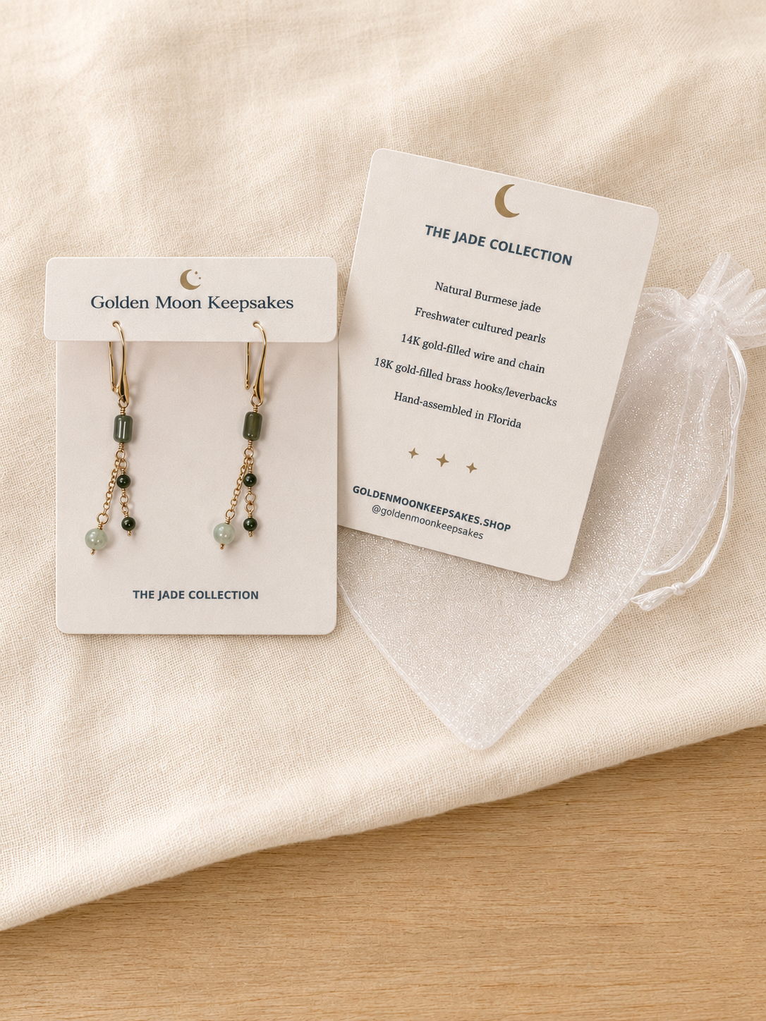 A pair of gold dangle earrings with green and pale blue beads, displayed on a white card labeled 'Golden Moon Keepsakes' alongside a white organza pouch and a product info card from 'The Jade Collection'. The cards are placed on a beige fabric surface over a light wood table.