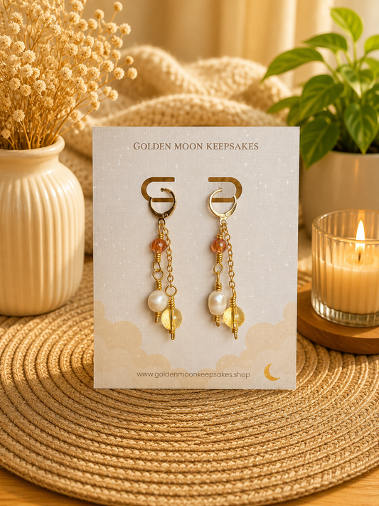 A pair of gold-toned dangle earrings displayed on a branded card, set on a woven placemat with dried flowers, a candle, and a plant in the background.