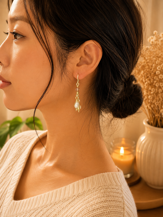 A woman in profile wearing a delicate gold dangle earring with a small clear faceted bead and a pale pearl-like drop, photographed in warm indoor lighting with a candle and dried flowers in the background.