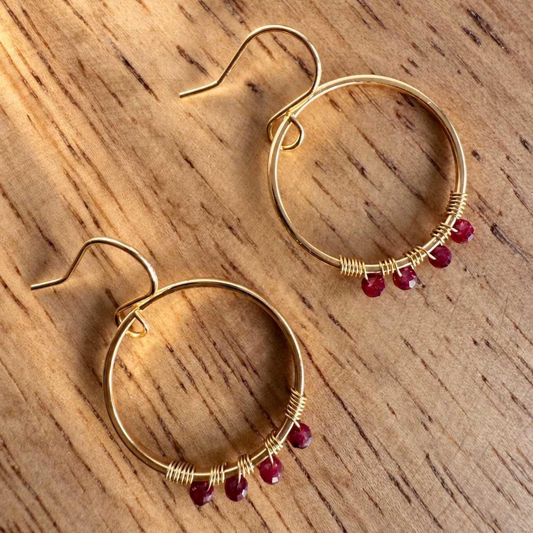 A pair of gold hoop earrings with small wrapped details and dangling red gemstone beads, displayed on a wooden surface.