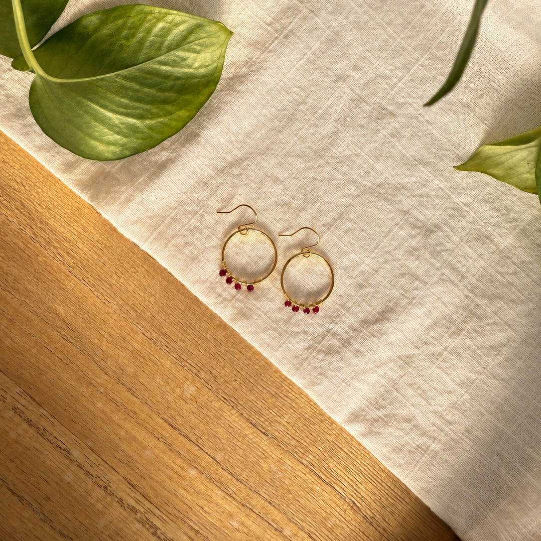 A pair of gold hoop earrings with small dangling red beads, displayed on a cream linen cloth over a wooden surface with green leaves in the corners.