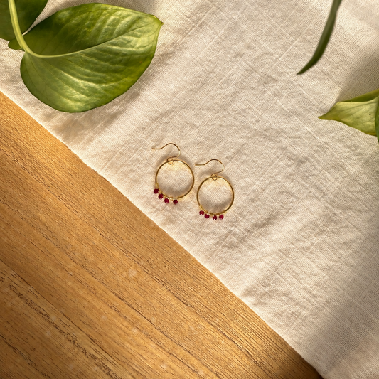 A pair of gold hoop earrings with small dangling red beads, displayed on a cream linen cloth over a wooden surface with green leaves in the corners.