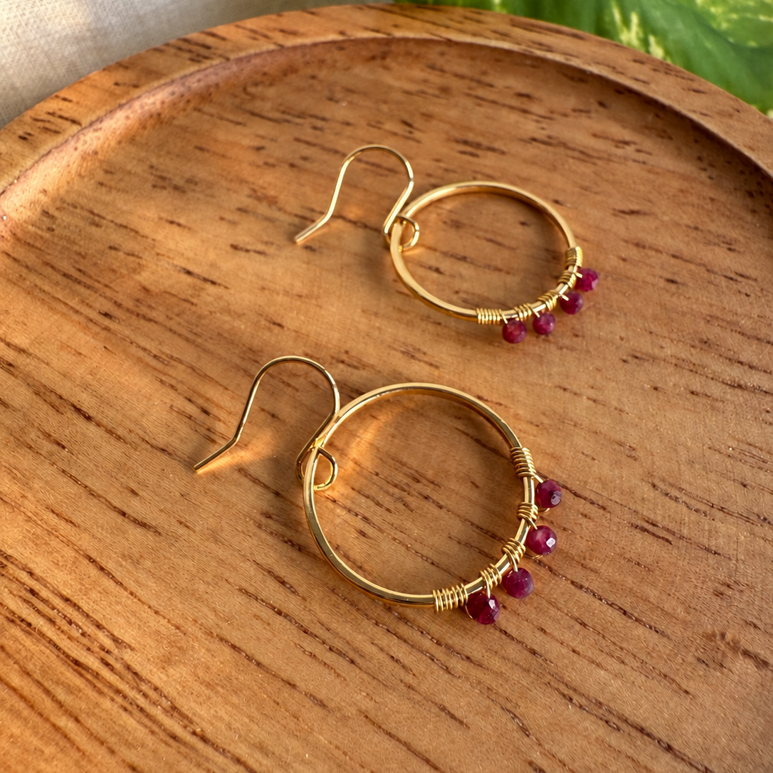 A pair of gold-tone hoop earrings with small wrapped details and tiny dark red beads at the bottom, resting on a round wooden tray.
