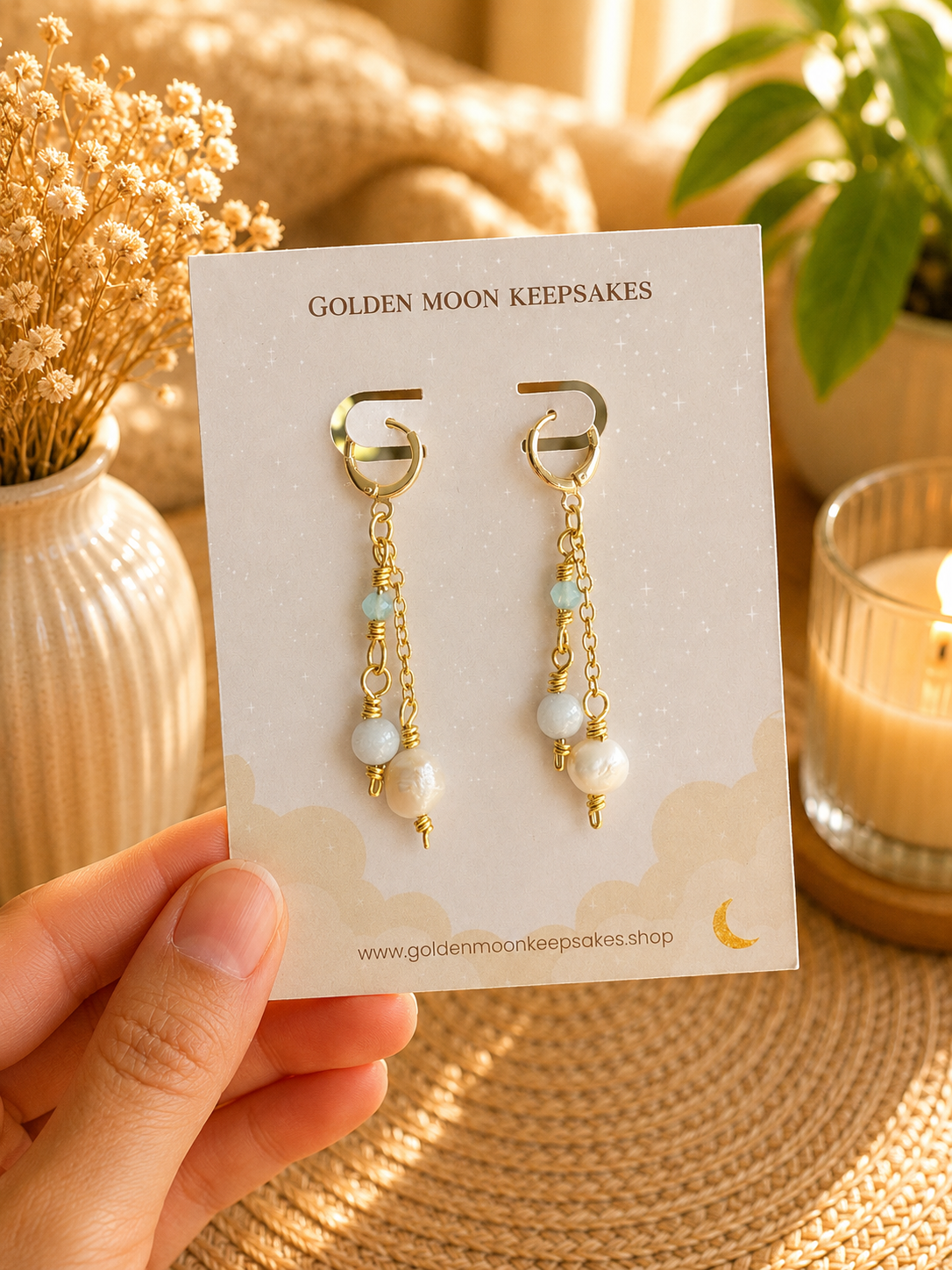 A hand holds a display card featuring a pair of dangling earrings with gold-tone hoops, small light blue beads, white pearl-like beads, and gold chain drops. The card reads 'GOLDEN MOON KEEPSAKES' and is photographed in a warm, styled setting with dried flowers, a candle, and a plant.