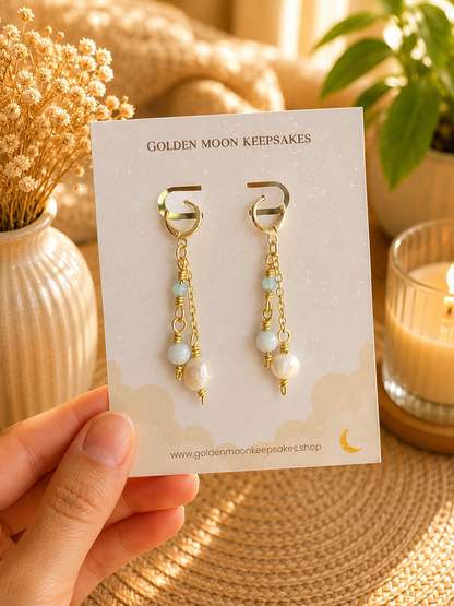 A hand holds a display card featuring a pair of dangling earrings with gold-tone hoops, small light blue beads, white pearl-like beads, and gold chain drops. The card reads 'GOLDEN MOON KEEPSAKES' and is photographed in a warm, styled setting with dried flowers, a candle, and a plant.