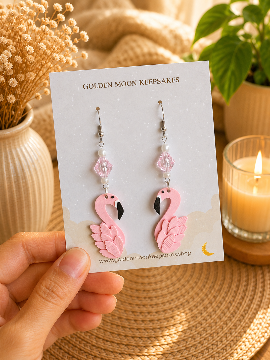 A hand holds a display card featuring a pair of pink flamingo dangle earrings with silver hooks and faceted pink beads. The card reads 'GOLDEN MOON KEEPSAKES' and is photographed in a warm, styled setting with a candle, dried flowers, and a plant in the background.