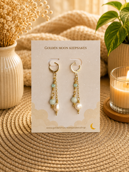 A pair of gold-tone dangle earrings with pale green beads and white pearls, displayed on a branded card. The card is styled on a woven mat with a candle, dried flowers, and a plant in the background.