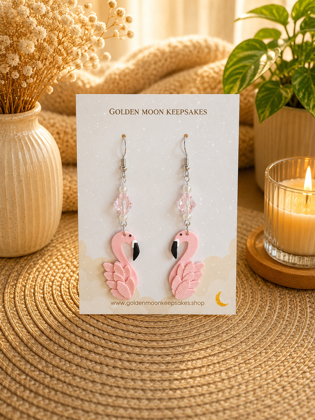 Pink flamingo dangle earrings displayed on a white card labeled 'GOLDEN MOON KEEPSAKES,' set on a woven mat with dried flowers, a candle, and a plant in the background.
